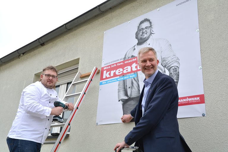 Marcus Ostendorf und Burghard Grupe bringen ein Plakat an die Häuserwand an.