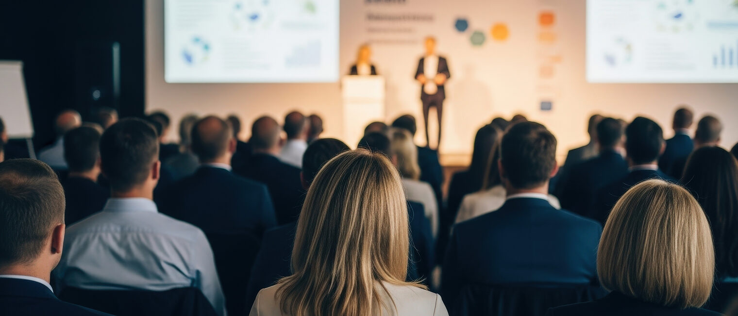 People listen at a business conference with speakers on stage and charts displayed